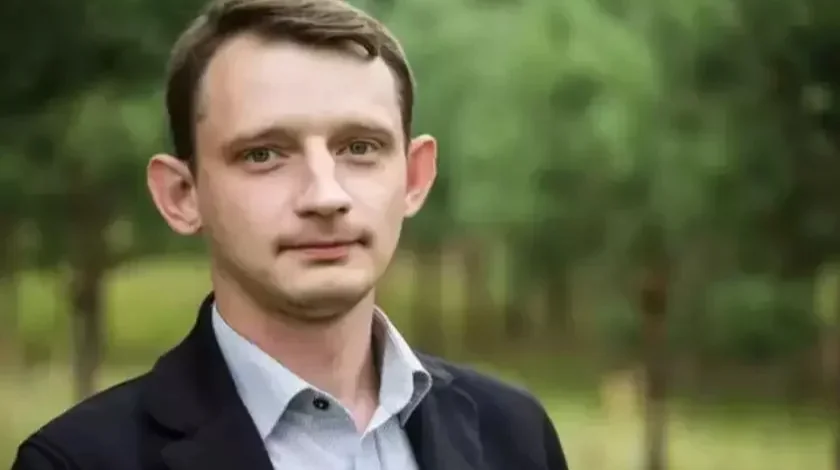 Молодой мужчина в светлой рубашке и тёмном пиджаке на фоне зелёных деревьев