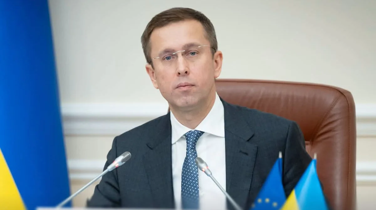 Мужчина в костюме и очках сидит за столом с микрофонами возле флагов Украины и ЕС