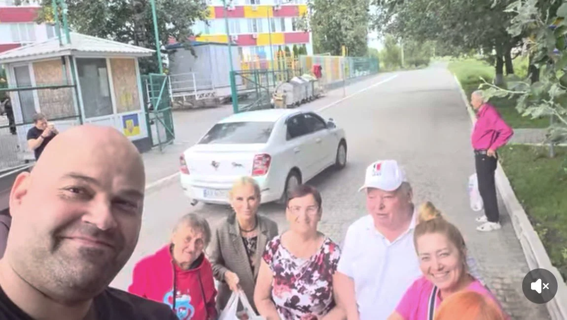 Лисий чоловік робить селфі з групою людей на вулиці біля білого авто