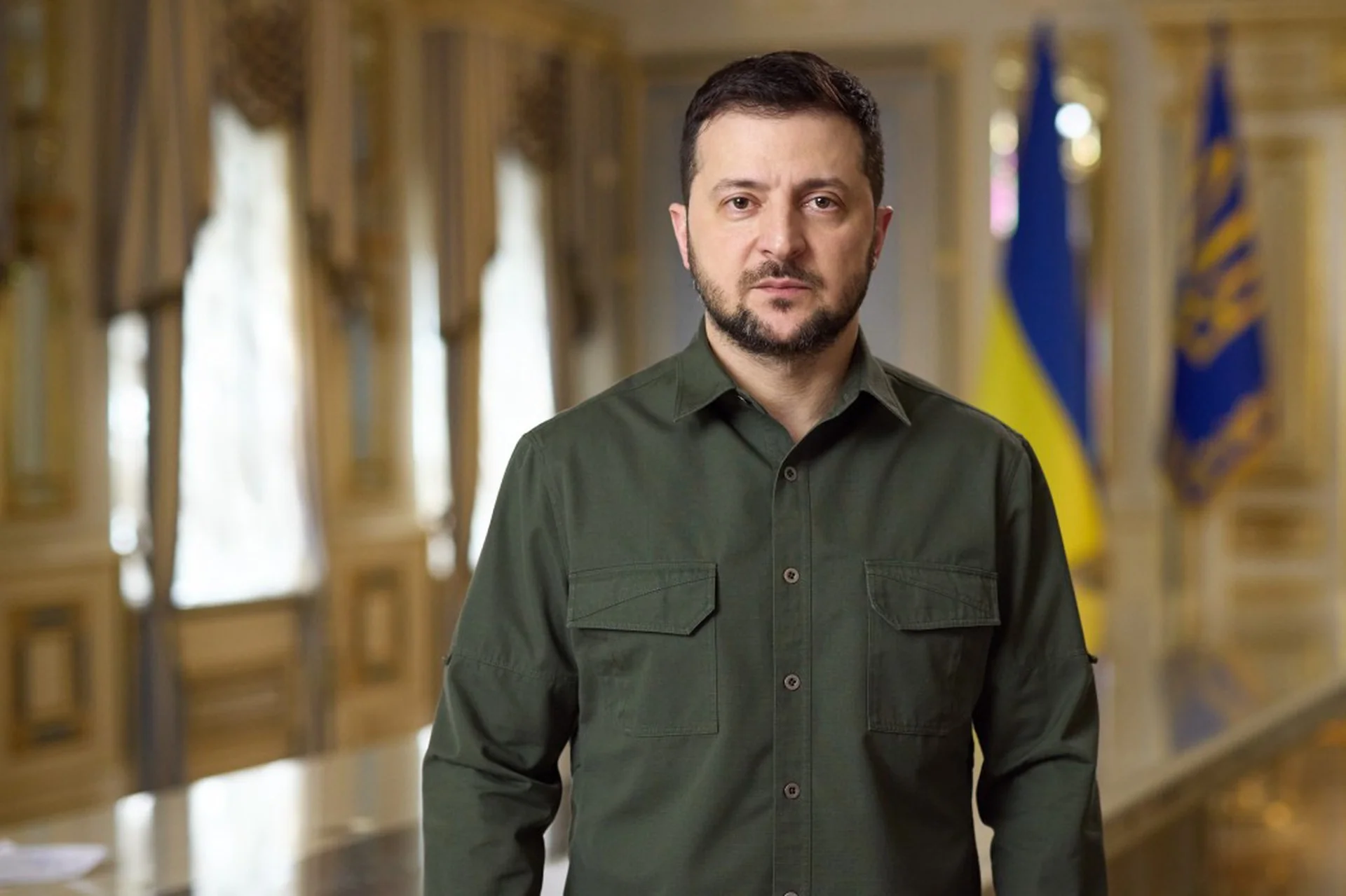 Володимир Зеленський у зеленій сорочці стоїть у залі з українськими прапорами