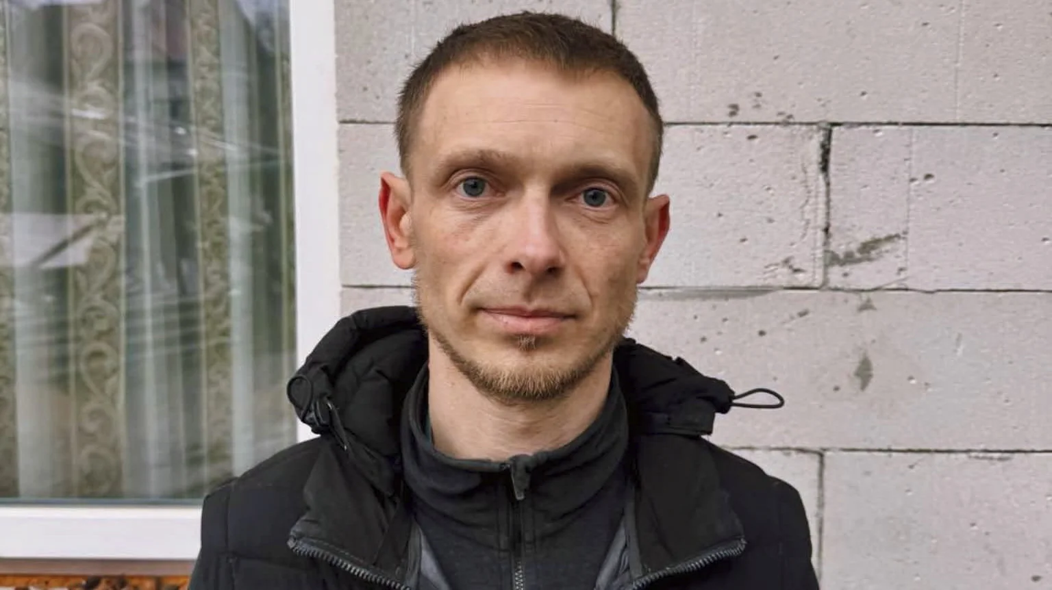 Чоловік у чорній куртці стоїть біля сірої цегляної стіни та вікна