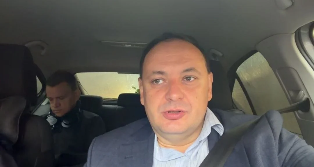 Чоловік у синьому піджаку говорить на камеру з салону авто