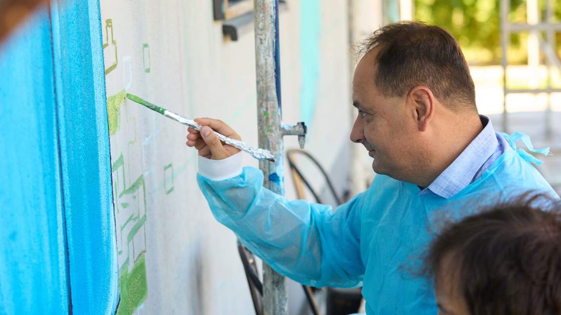 Чоловік у синій куртці малює на стіні пензлем, створюючи мистецький мурал