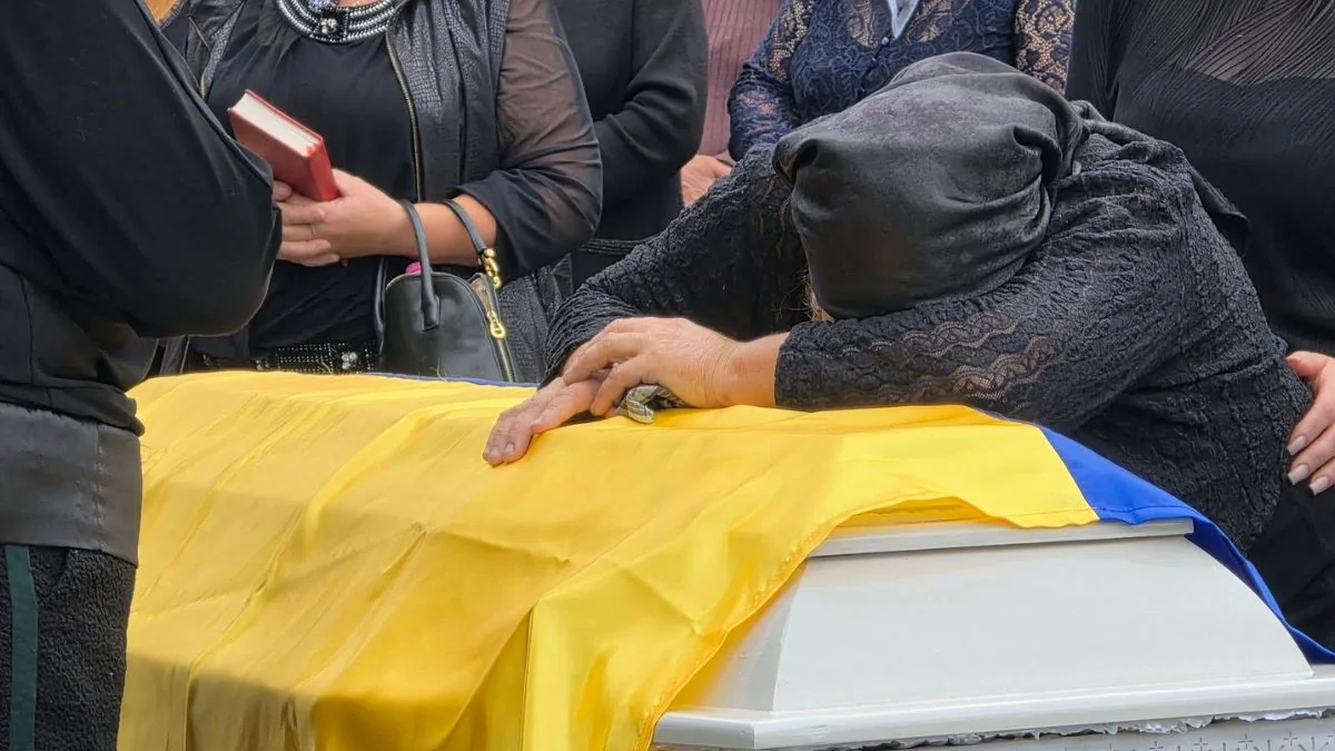 Жінка в чорному схилилася над труною, накритою жовтим українським прапором